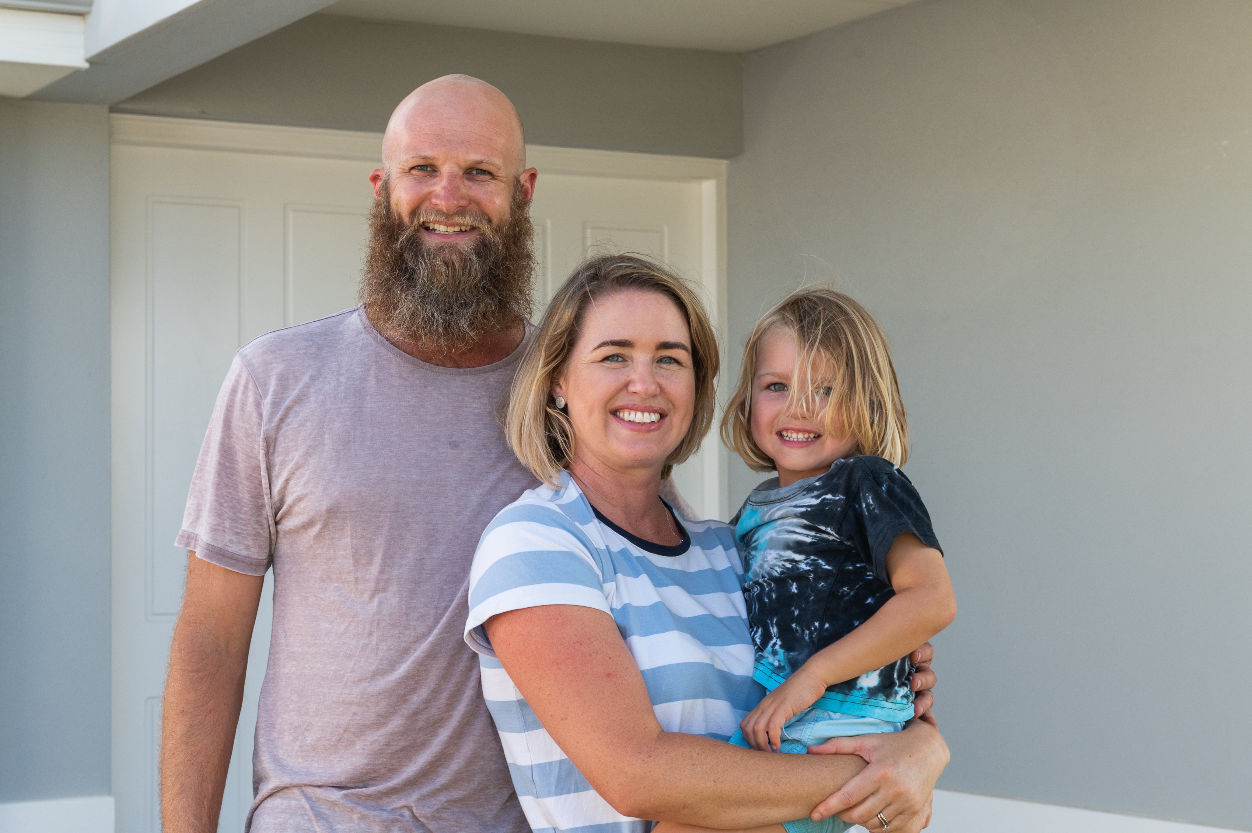 Smiling bearded bald man next to smiling blonde woman holding smiling blonde child in front of white front door. 
