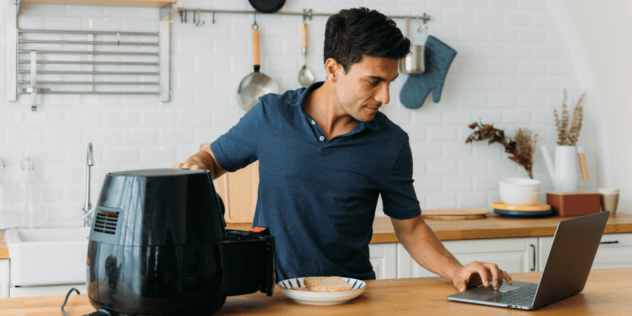 Man in home kitchen using air-fryer and laptop. 
