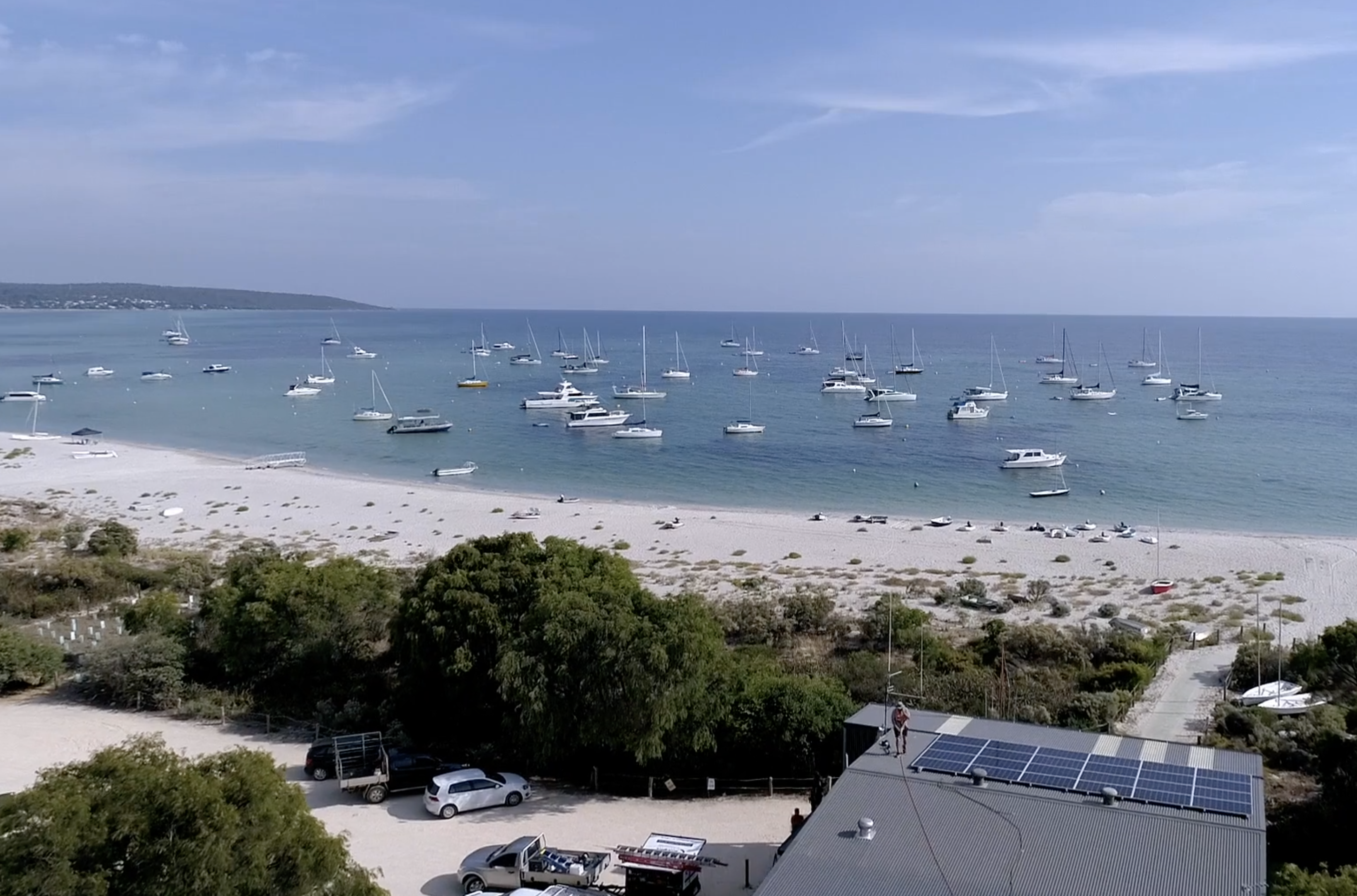 Beach on a sunny day with boats in the water and a grey shack with solar panels being installed on the roof by a man Beach on a sunny day with boats in the water and a grey shack with solar panels being installed on the roof by a man
