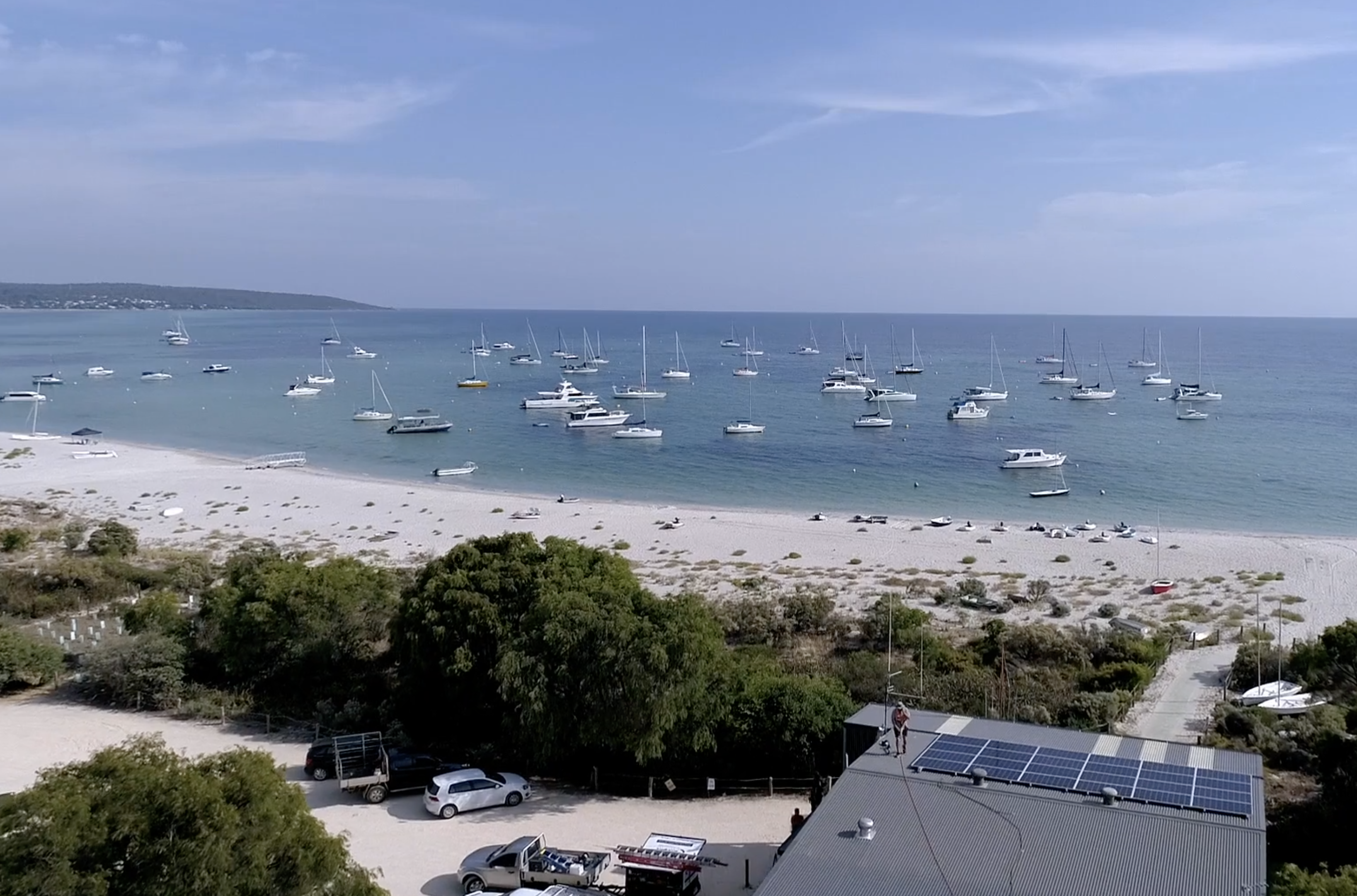 Beach on a sunny day with boats in the water and a grey shack with solar panels being installed on the roof by a man